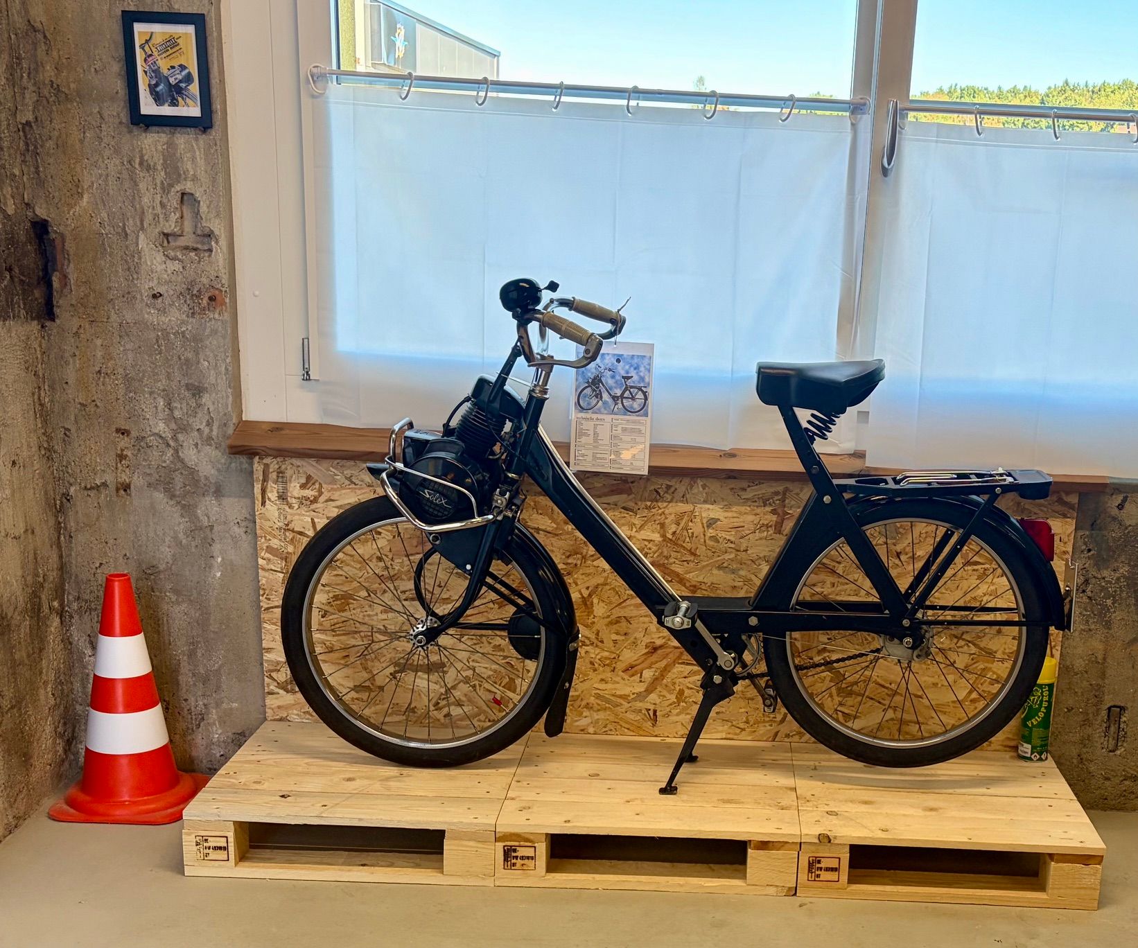 Ein umgestaltetes, schwarzes Solex-Mofa steht auf einer Holzpalette im Atelier.